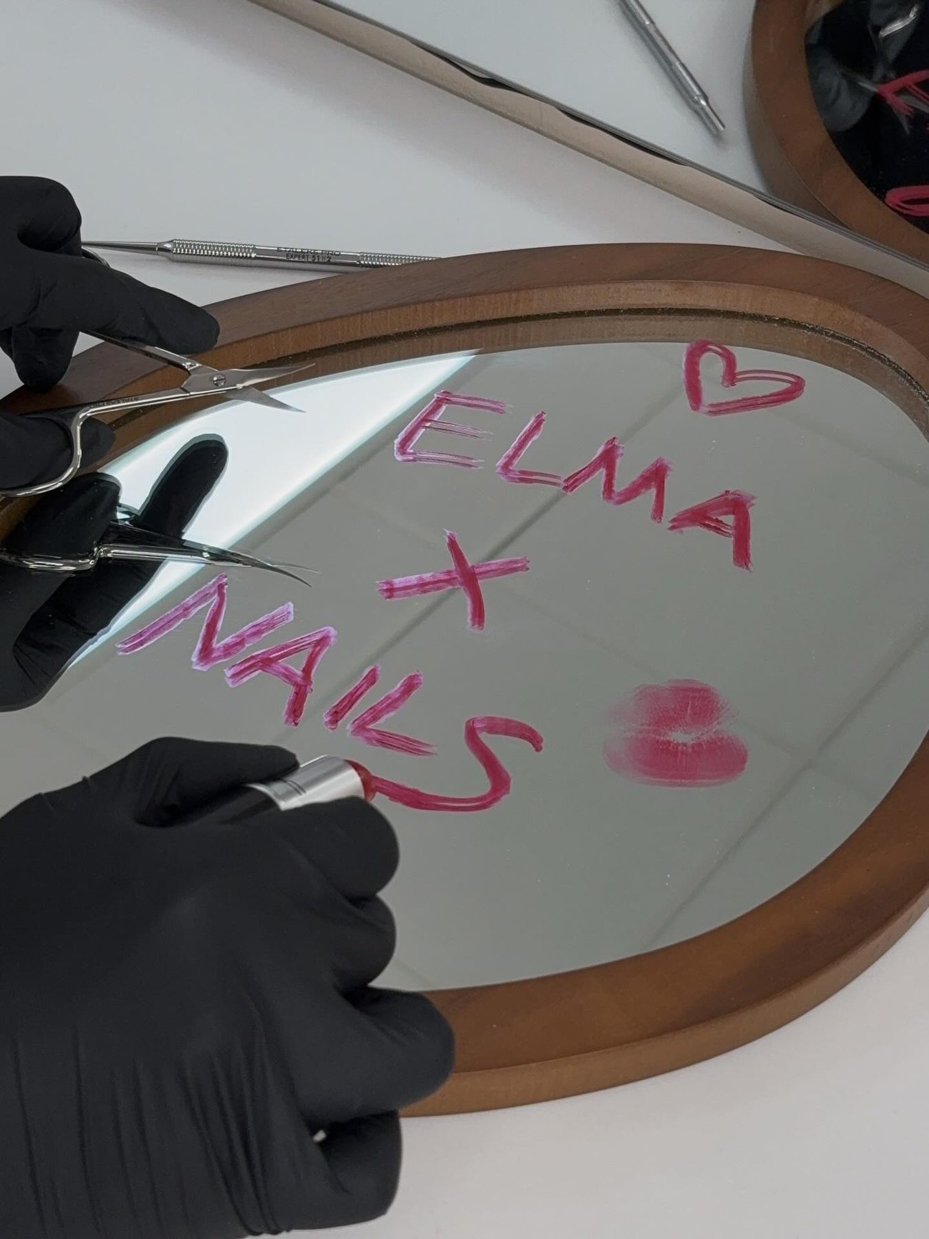 Ronde spiegel met ELMA x NAILS in lippenstift, handen in zwarte handschoenen met nagelriemtang